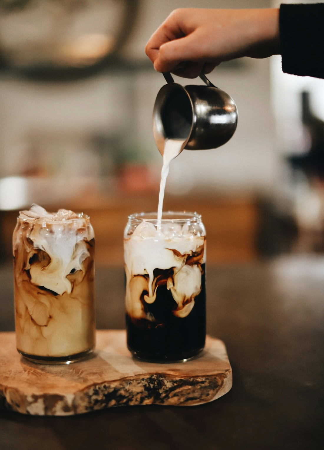 A hand pouring cream into a glass of iced coffee on a wooden board