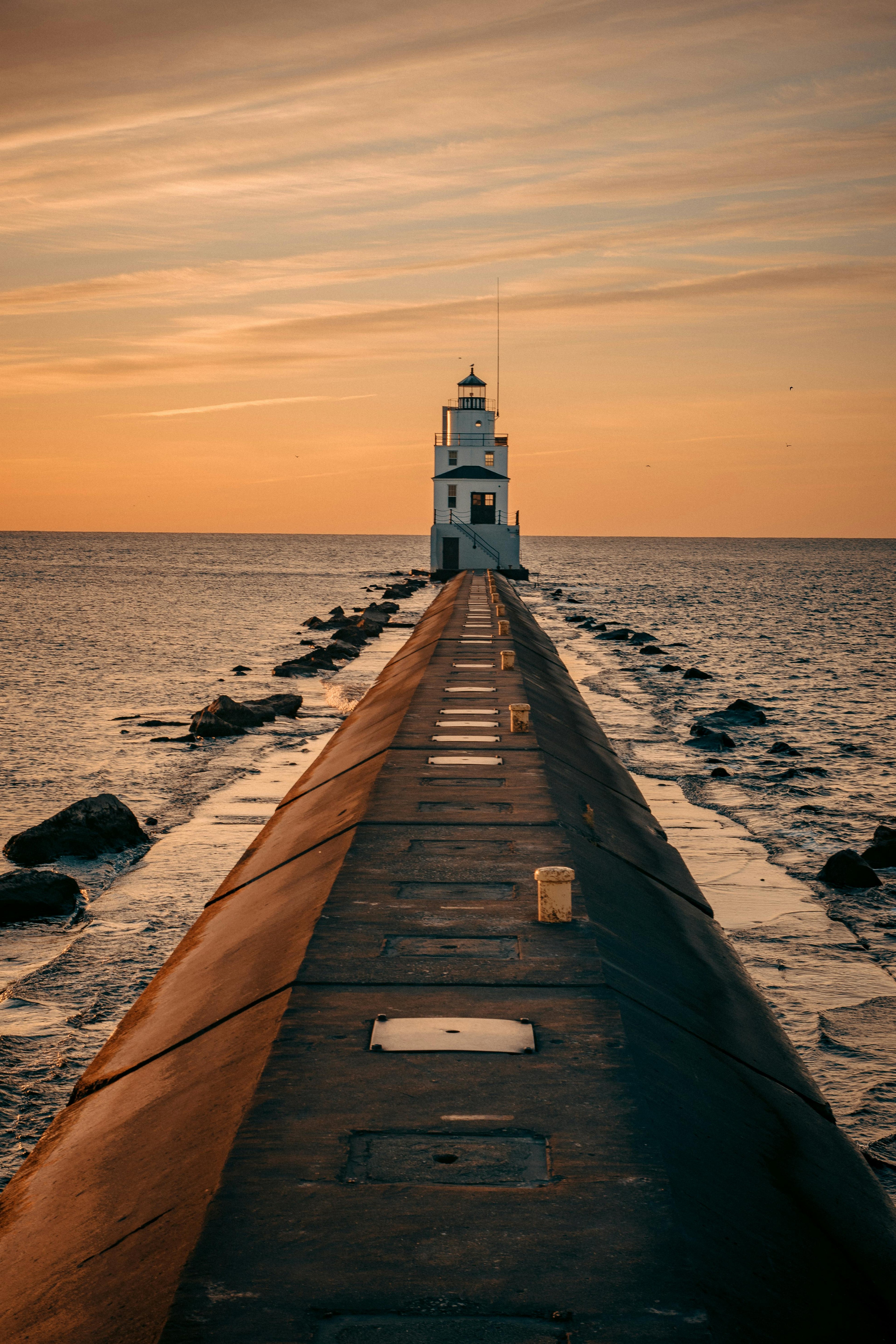 The St. Joseph North Pier Lighthouse stands at the end of a long pier extending into calm waters. The pier is lined with yellow bollards and rectangular covers. The sky is painted with warm hues of orange and yellow, indicating sunrise or sunset. The scene is tranquil and picturesque
